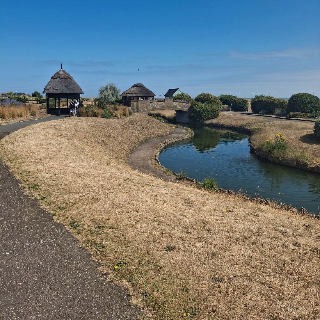 The Venetian Waterways at Great Yarmouth, a winding green canal curving past thatched shelters and a small stone bridge